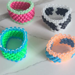 May include: Four colorful beaded rings, each with a different color combination. The rings are made of small, round beads in shades of pink, green, blue, and black. The rings are arranged on a white surface.