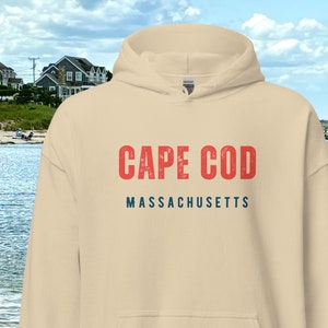 May include: A cream-coloured hoodie featuring the text "CAPE COD" in red and "MASSACHUSETTS" in blue. The sweatshirt has a front pocket and a drawstring hood. The background shows a coastal scene with houses and water.