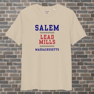 Può includere: Una maglietta beige con una stampa grafica blu e rossa che recita "SALEM LEAD MILLS MASSACHUSETTS".