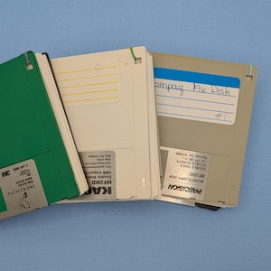 May include: A collection of 3.5-inch floppy disks in green, white, and beige. The disks have labels with text including "IMATION", "PRECISION", and "Compaq Fix Disk". The green disk is labeled "1.44 MB 3M".