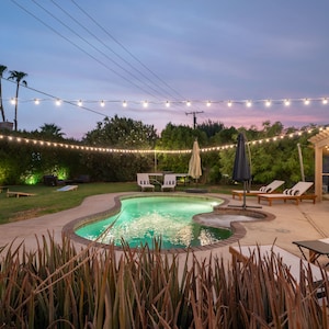 May include: A swimming pool with a curved edge and a patio with lounge chairs and a pergola. String lights are strung across the yard, creating a festive atmosphere.