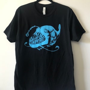 Op de afbeelding: Een zwart T-shirt met een blauwe anglerfish-print. De vis heeft een grote bek met scherpe tanden en een bioluminescent lokmiddel.