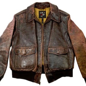 Könnte beinhalten: Eine braune Lederbomberjacke mit Reißverschluss und zwei Pattentaschen. Die Jacke hat einen Used-Look und ein gelbes Futter mit einem schwarzen und roten Etikett, auf dem "Let's Do It" steht.