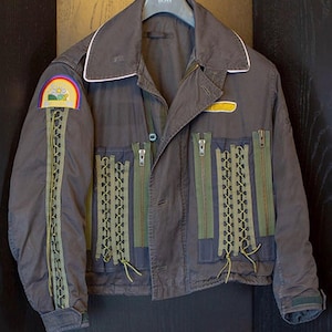 May include: A dark grey jacket with a white collar and green zipper details. The jacket has a patch on the left sleeve with a rainbow and a sun. The jacket also has a yellow patch on the right side.