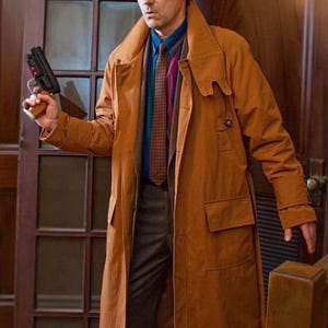 Könnte beinhalten: Ein Mann in einem braunen Trenchcoat und einer gemusterten Krawatte hält eine schwarze Waffe in seiner rechten Hand. Er steht in einem Durchgang mit einer Holztür und einem Glasfenster.