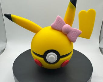May include: A yellow and black Pikachu-themed decorative item with a pink bow. The item has a round yellow body with black accents, red circles, and a white button. The ears and tail tips are black.