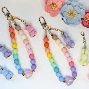 May include: Rainbow beaded keychains with gold-tone hardware. Each keychain features a gradient of colorful beads, a heart charm, and a small animal charm, including bears and bunnies. The background includes pink and blue floral accents.
