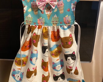 May include: A white kitchen towel with a pattern of cats wearing hats and holding fish. The towel is trimmed with teal lace and has a blue and white ice cream cone pattern on the top. A pink bow with white polka dots is attached to the top of the towel.