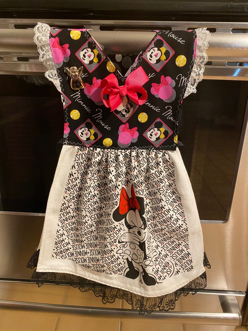 Puede incluir: Una toalla de cocina en blanco y negro con un dise&ntilde;o de Minnie Mouse. La toalla tiene un lazo rosa y un borde de encaje blanco y negro. La toalla est&aacute; colgada en una puerta de horno de plata.