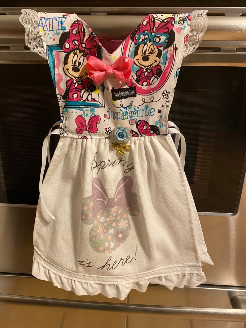 May include: White kitchen towel with a pink and white Minnie Mouse design. The towel has a ruffle trim and says "Spring is here!" in black text. The towel is hanging on a silver oven door.
