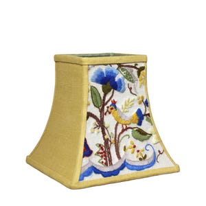May include: A yellow lampshade with an embroidered floral design featuring blue flowers, green leaves, and yellow birds. The lampshade has a square shape with rounded corners.
