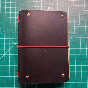 Op de afbeelding: Een zwart leren notitieboek met een rode elastische sluiting. Het notitieboek is vastgezet met vier messing klinknagels en rode stiksels langs de rug. De pagina's zijn zichtbaar langs de rand. Het notitieboek ligt op een groene snijmat.