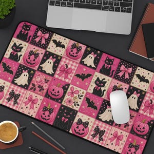 May include: A rectangular desk mat featuring a repeating patchwork pattern of pink and black Halloween motifs, including jack-o'-lanterns, black cats, ghosts, bats, and bows, set against a background of pink gingham and floral accents.