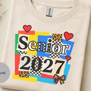 以下が含まれることがあります： 「Senior」の文字と「2027」の数字がコラージュデザインでプリントされたクリーム色のTシャツ。デザインには赤いハート、リボン、ヒョウ柄のアクセントが含まれています。このシャツは柔らかい素材で作られています。