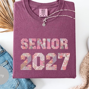 Op de afbeelding: Een bessenkleurig T-shirt met de tekst "SENIOR 2027" in een bloemen patchwork ontwerp. Het shirt wordt gecombineerd met een blauwe jeans, een zilveren ketting en witte schoenen. Het shirt heeft een ronde hals en korte mouwen.