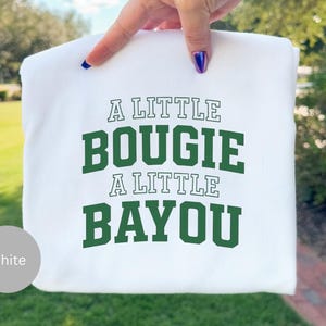 May include: White sweatshirt with the text "A LITTLE BOUIGIE A LITTLE BAYOU" in green block letters. The sweatshirt is held up by a hand with a gold bracelet and blue nail polish. The background is blurred green foliage.