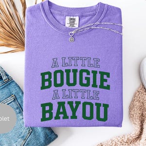 Puede incluir: Una sudadera violeta con el texto "A LITTLE BOUGIE A LITTLE BAYOU" en verde. Un collar plateado con un colgante adorna la prenda. La imagen incluye vaqueros azules y zapatos blancos sin cordones.