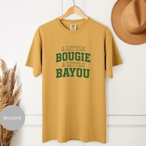 Puede incluir: Camiseta de color mostaza con el texto "A Little Bougie A Little Bayou" en letras verdes. La camiseta est&aacute; colgada de una percha de madera. Un sombrero marr&oacute;n es visible en el fondo.