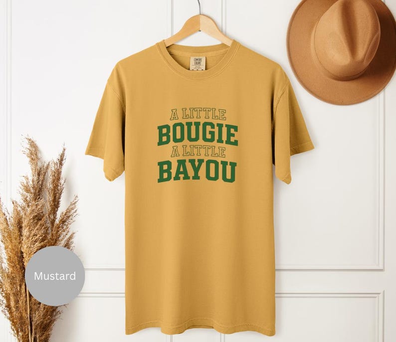 Puede incluir: Camiseta de color mostaza con el texto "A Little Bougie A Little Bayou" en letras verdes. La camiseta est&aacute; colgada de una percha de madera. Un sombrero marr&oacute;n es visible en el fondo.