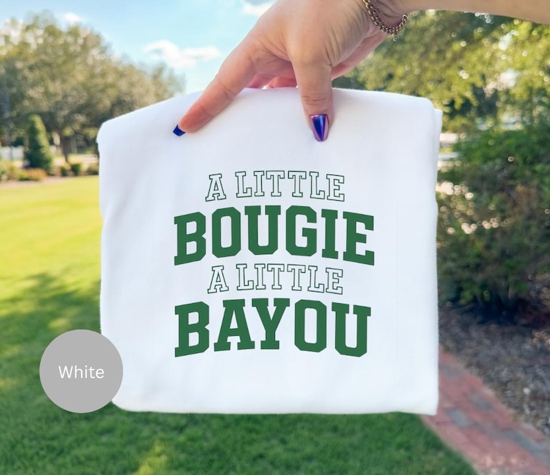 May include: White sweatshirt with the text "A LITTLE BOUIGIE A LITTLE BAYOU" in green block letters. The sweatshirt is held up by a hand with a gold bracelet and blue nail polish. The background is blurred green foliage.