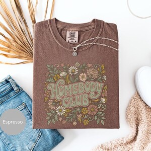 Puede incluir: Camiseta color espresso con el texto "Homebody Club" en una fuente retro, rodeada de ilustraciones florales en tonos verdes, rosas y crema. Un collar plateado con un colgante descansa sobre la camiseta. La camiseta se combina con vaqueros azules y zapatillas blancas.