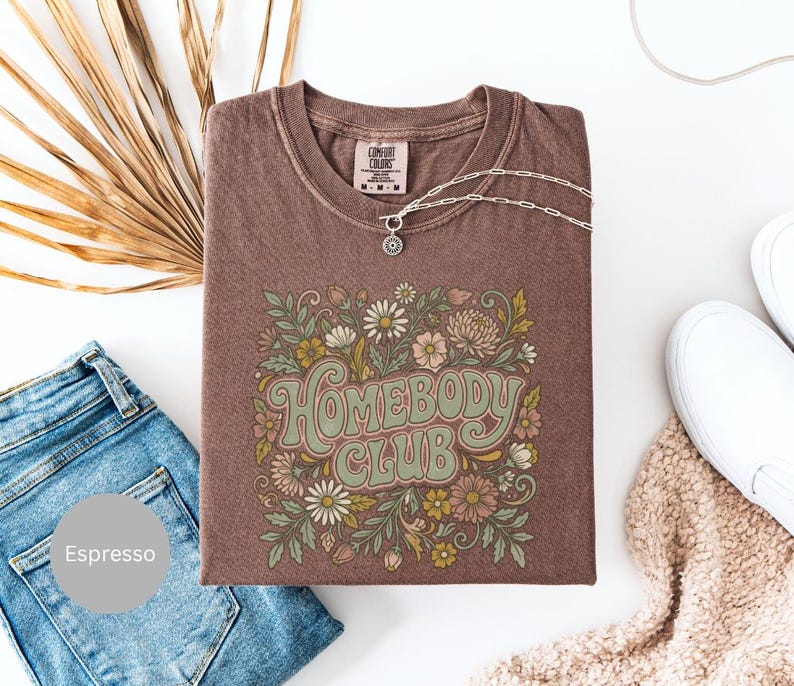 Puede incluir: Camiseta color espresso con el texto "Homebody Club" en una fuente retro, rodeada de ilustraciones florales en tonos verdes, rosas y crema. Un collar plateado con un colgante descansa sobre la camiseta. La camiseta se combina con vaqueros azules y zapatillas blancas.
