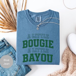 Puede incluir: Una camiseta azul con el texto "A Little Bougie A Little Bayou" en verde. La camiseta est&aacute; doblada y se muestra con vaqueros azules, un collar plateado y zapatos blancos.