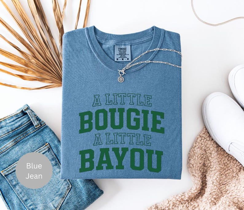 Puede incluir: Una camiseta azul con el texto "A Little Bougie A Little Bayou" en verde. La camiseta est&aacute; doblada y se muestra con vaqueros azules, un collar plateado y zapatos blancos.