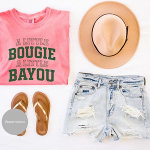 Puede incluir: Una presentaci&oacute;n plana de ropa de verano que incluye una camiseta rosa con el texto "A Little Bougie A Little Bayou", pantalones cortos de mezclilla de lavado claro, un sombrero color canela y sandalias marrones con correas doradas. Las sandalias est&aacute;n junto a un c&iacute;rculo gris con la palabra "Watermelon".