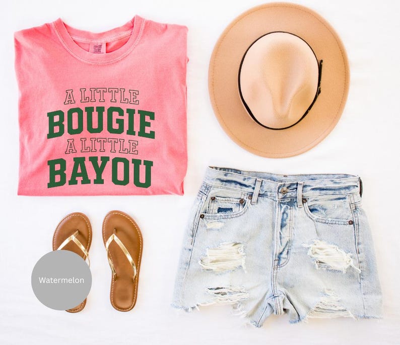 Puede incluir: Una presentaci&oacute;n plana de ropa de verano que incluye una camiseta rosa con el texto "A Little Bougie A Little Bayou", pantalones cortos de mezclilla de lavado claro, un sombrero color canela y sandalias marrones con correas doradas. Las sandalias est&aacute;n junto a un c&iacute;rculo gris con la palabra "Watermelon".