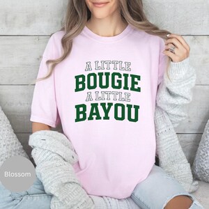 Puede incluir: Camiseta rosa claro con el texto "A LITTLE BOUGIE A LITTLE BAYOU" en letras verdes y blancas. La camiseta es de estilo casual, adecuada para el uso diario.