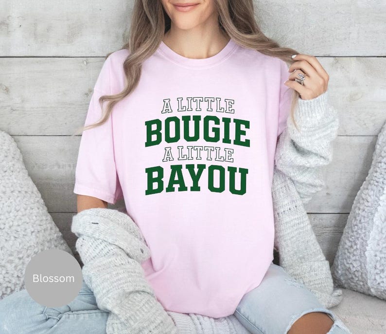 Puede incluir: Camiseta rosa claro con el texto "A LITTLE BOUGIE A LITTLE BAYOU" en letras verdes y blancas. La camiseta es de estilo casual, adecuada para el uso diario.