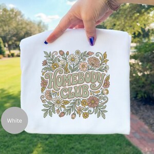 Puede incluir: Sudadera blanca con la inscripci&oacute;n "Homebody Club" en una fuente retro, rodeada de un dise&ntilde;o floral. El dise&ntilde;o presenta margaritas, crisantemos y hojas en tonos verdes, melocot&oacute;n y marr&oacute;n. La sudadera es sostenida por una mano.