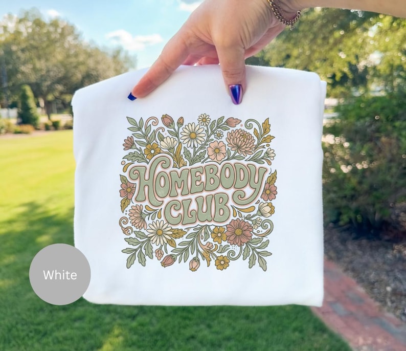 Puede incluir: Sudadera blanca con la inscripci&oacute;n "Homebody Club" en una fuente retro, rodeada de un dise&ntilde;o floral. El dise&ntilde;o presenta margaritas, crisantemos y hojas en tonos verdes, melocot&oacute;n y marr&oacute;n. La sudadera es sostenida por una mano.