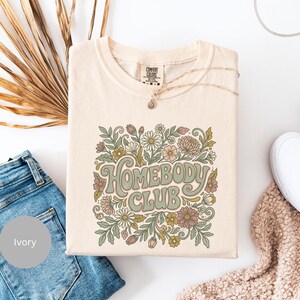 Puede incluir: Camiseta color marfil con la frase "Homebody Club" en una fuente retro, rodeada de un dise&ntilde;o floral en tonos verdes, rosas y amarillos. La camiseta se combina con vaqueros azules, un collar y zapatos blancos.