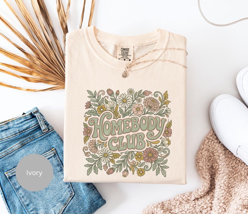 Puede incluir: Camiseta color marfil con la frase "Homebody Club" en una fuente retro, rodeada de un dise&ntilde;o floral en tonos verdes, rosas y amarillos. La camiseta se combina con vaqueros azules, un collar y zapatos blancos.