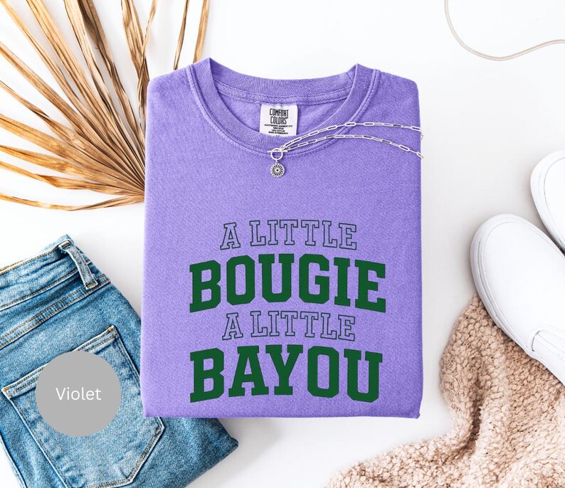 Puede incluir: Una sudadera violeta con el texto "A LITTLE BOUGIE A LITTLE BAYOU" en verde. Un collar plateado con un colgante adorna la prenda. La imagen incluye vaqueros azules y zapatos blancos sin cordones.