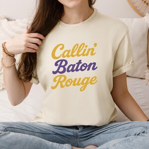 Puede incluir: Camiseta color crema con el texto "Callin' Baton Rouge" en escritura amarilla y morada. La camiseta tiene mangas cortas y un corte relajado. El texto está apilado verticalmente.