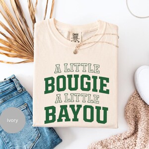 Puede incluir: Camiseta de color marfil con el texto "A LITTLE BOUGIE A LITTLE BAYOU" en verde y blanco. La camiseta est&aacute; doblada y colocada sobre una superficie blanca, junto a unos vaqueros azules y zapatos blancos. Tambi&eacute;n se ve un collar y una planta seca.