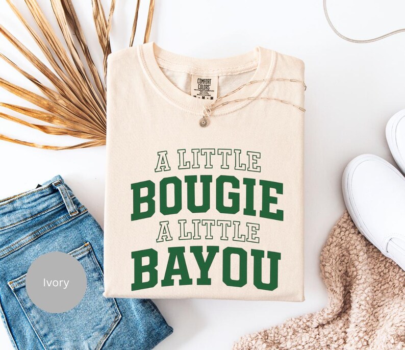 Puede incluir: Camiseta de color marfil con el texto "A LITTLE BOUGIE A LITTLE BAYOU" en verde y blanco. La camiseta est&aacute; doblada y colocada sobre una superficie blanca, junto a unos vaqueros azules y zapatos blancos. Tambi&eacute;n se ve un collar y una planta seca.