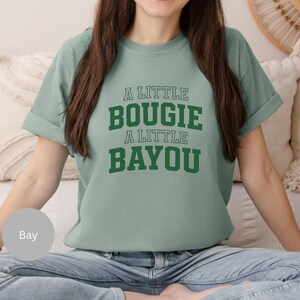 Puede incluir: Una camiseta verde claro con el texto "A LITTLE BOUGIE A LITTLE BAYOU" en letras verdes. La camiseta tiene mangas cortas y un corte relajado. La modelo lleva vaqueros azules.