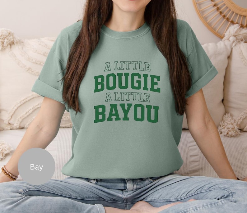 Puede incluir: Una camiseta verde claro con el texto "A LITTLE BOUGIE A LITTLE BAYOU" en letras verdes. La camiseta tiene mangas cortas y un corte relajado. La modelo lleva vaqueros azules.