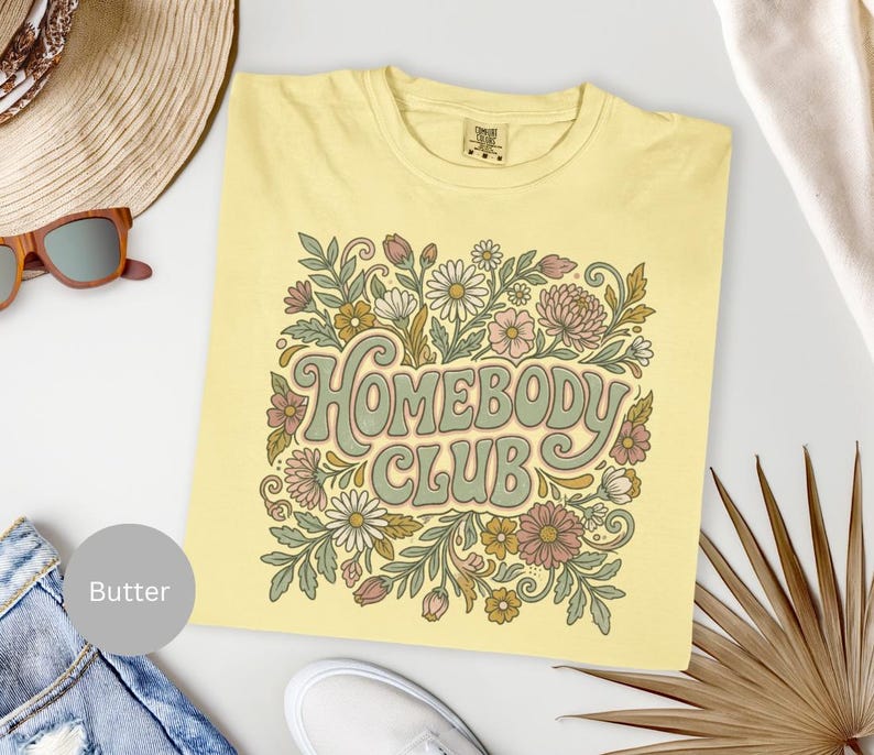 Puede incluir: Camiseta de color mantequilla con un dise&ntilde;o floral y las palabras "Homebody Club" en una fuente retro. El dise&ntilde;o presenta margaritas, hojas y flores en tonos de rosa, verde y marr&oacute;n. La camiseta est&aacute; doblada y expuesta sobre una superficie blanca.