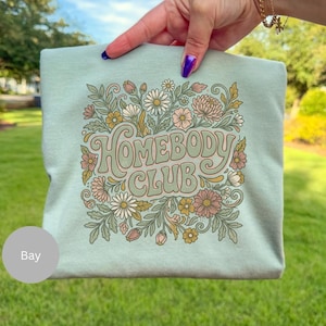 Puede incluir: Sudadera verde claro con la frase "HOMEBODY CLUB" en tipograf&iacute;a retro, rodeada de un dise&ntilde;o floral en tonos rosas, blancos y verdes. La sudadera es sostenida por una mano, con un fondo de hierba.