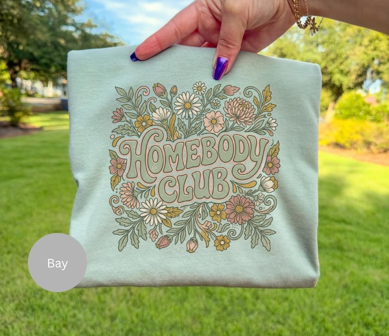 Puede incluir: Sudadera verde claro con la frase "HOMEBODY CLUB" en tipograf&iacute;a retro, rodeada de un dise&ntilde;o floral en tonos rosas, blancos y verdes. La sudadera es sostenida por una mano, con un fondo de hierba.