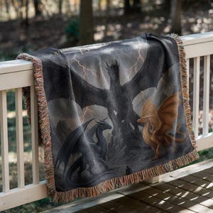 May include: A woven throw blanket featuring a fantasy dragon scene in shades of gray, black, and gold. The design includes multiple dragons, lightning bolts, and a dark, stormy sky. The blanket has a fringed edge and is draped over a white railing.