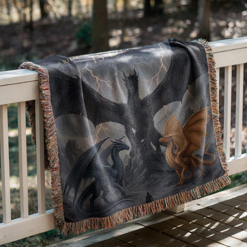 May include: A woven throw blanket featuring a fantasy dragon scene in shades of gray, black, and gold. The design includes multiple dragons, lightning bolts, and a dark, stormy sky. The blanket has a fringed edge and is draped over a white railing.
