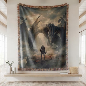 May include: Woven tapestry throw blanket featuring a fantasy scene with a large dragon and a figure in a dark coat. The artwork is in shades of gray, black, and brown, with a fringed border. The blanket is displayed on a wall.