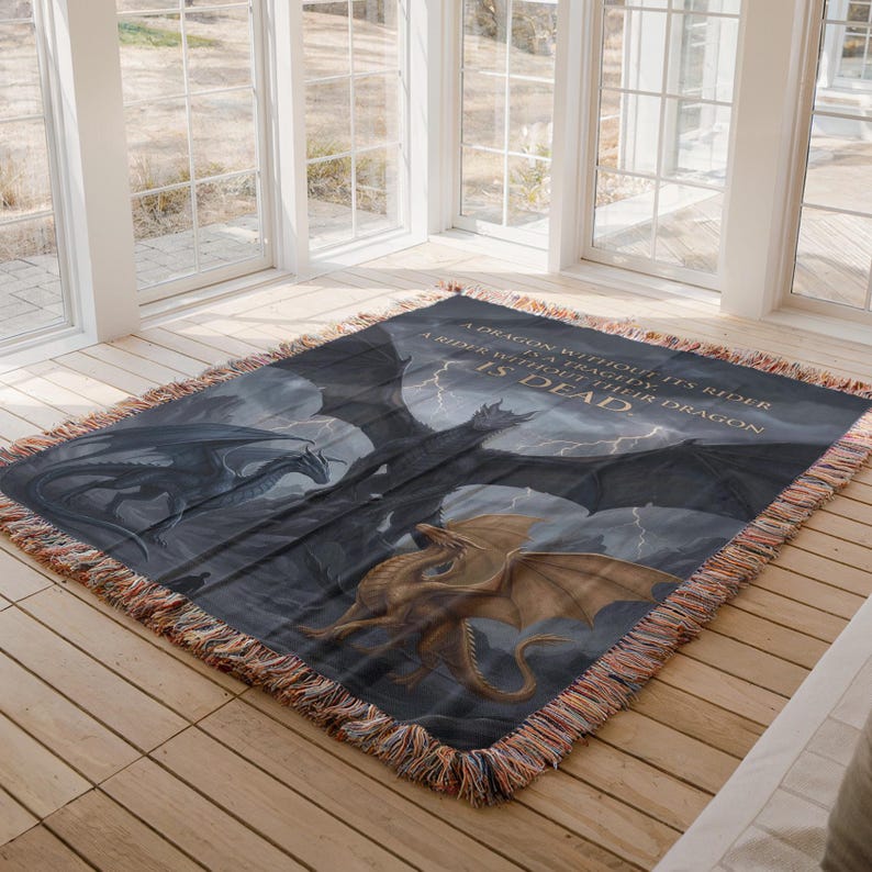 May include: A woven throw blanket featuring a fantasy dragon scene with a quote: "A dragon without its rider is a tragedy, a rider without their dragon is dead." The blanket has a dark blue background with illustrations of dragons in black and gold.