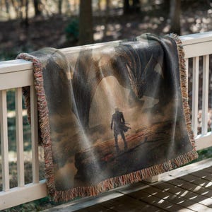 May include: Woven throw blanket featuring a fantasy scene with a dragon and a figure in a long coat. The blanket has a fringed edge and is draped over a white railing. The color palette includes shades of brown, gray, and orange.